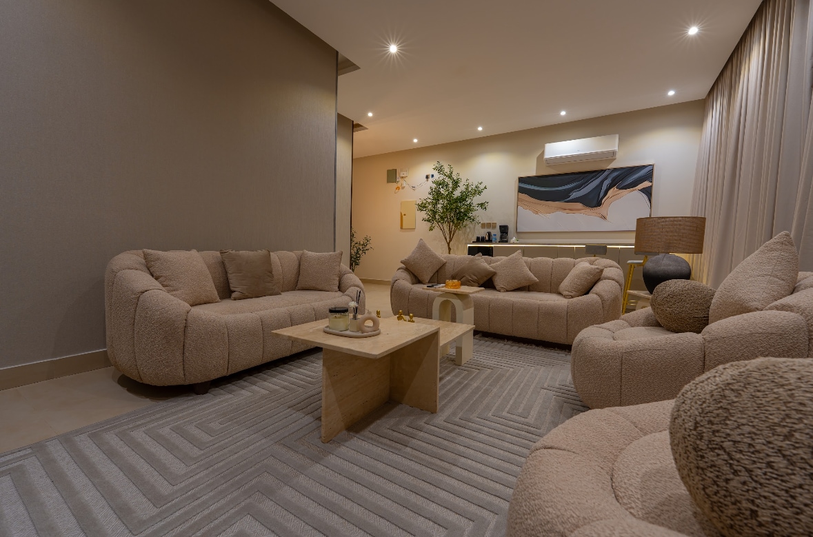 A spacious living area features soft, plush sofas arranged around a modern wooden coffee table. Warm lighting enhances the neutral color palette, while an abstract artwork adorns the wall. A small plant adds a touch of greenery and warmth to the space.