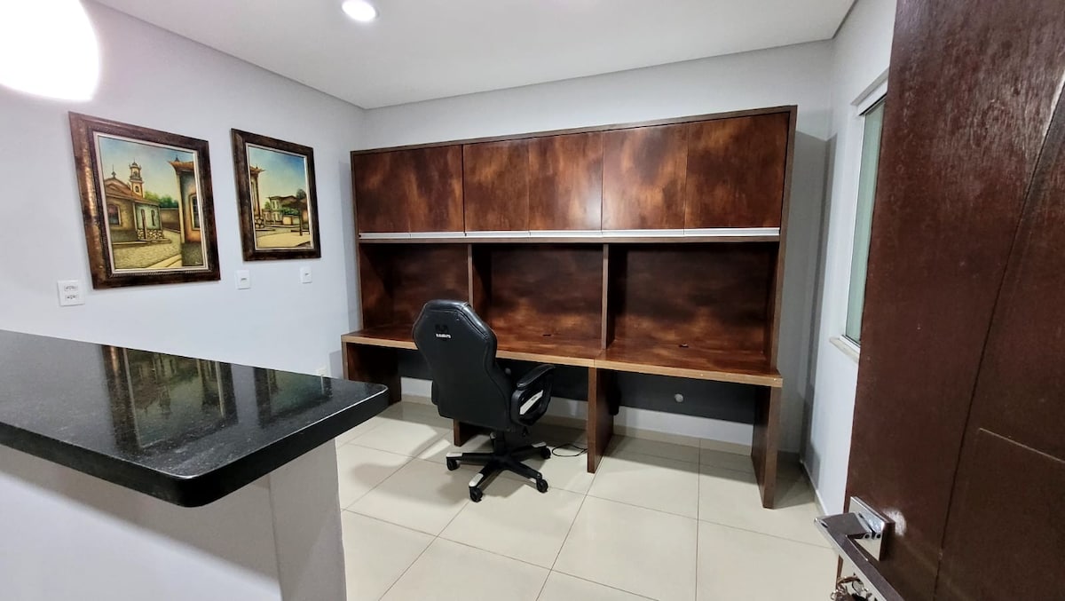 A functional workspace features a large wooden desk with an upper storage unit. A black rolling chair is positioned in front of the desk. Two framed paintings are displayed on the wall, providing a decorative element to the room. Natural light is provided by a side window.