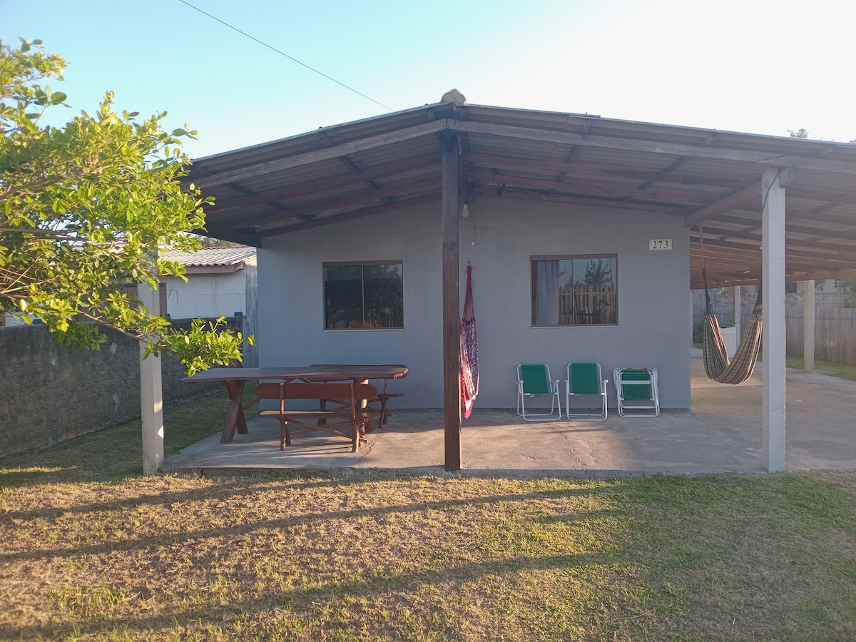 A simple outdoor area is presented, featuring a covered patio with a wooden table and several chairs. Two hammocks are visible, hanging nearby under the shelter. The ground is grassy, and walls of the house are painted in a neutral color, with large windows providing natural light.