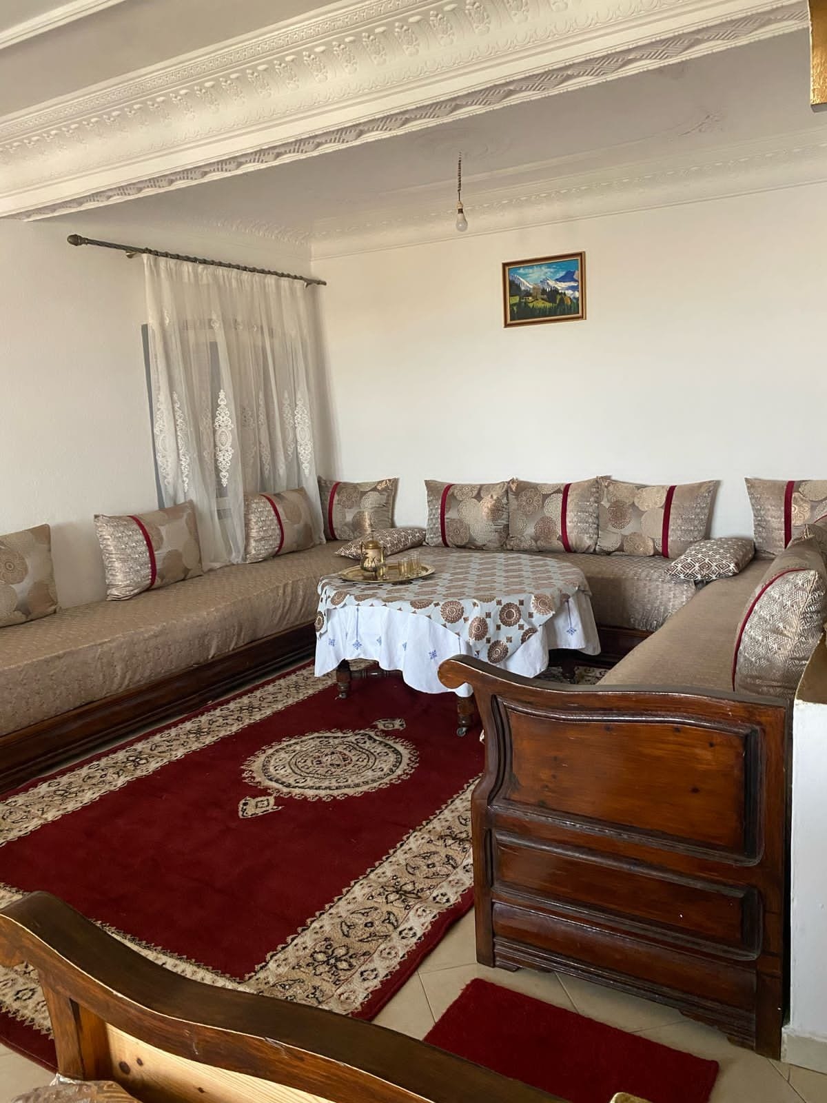 A welcoming living area is furnished with a plush seating arrangement, featuring patterned cushions. A small table is set at the center, adorned with a decorative cloth and traditional items. Light-colored curtains frame a window, while a piece of art adds character to the wall.