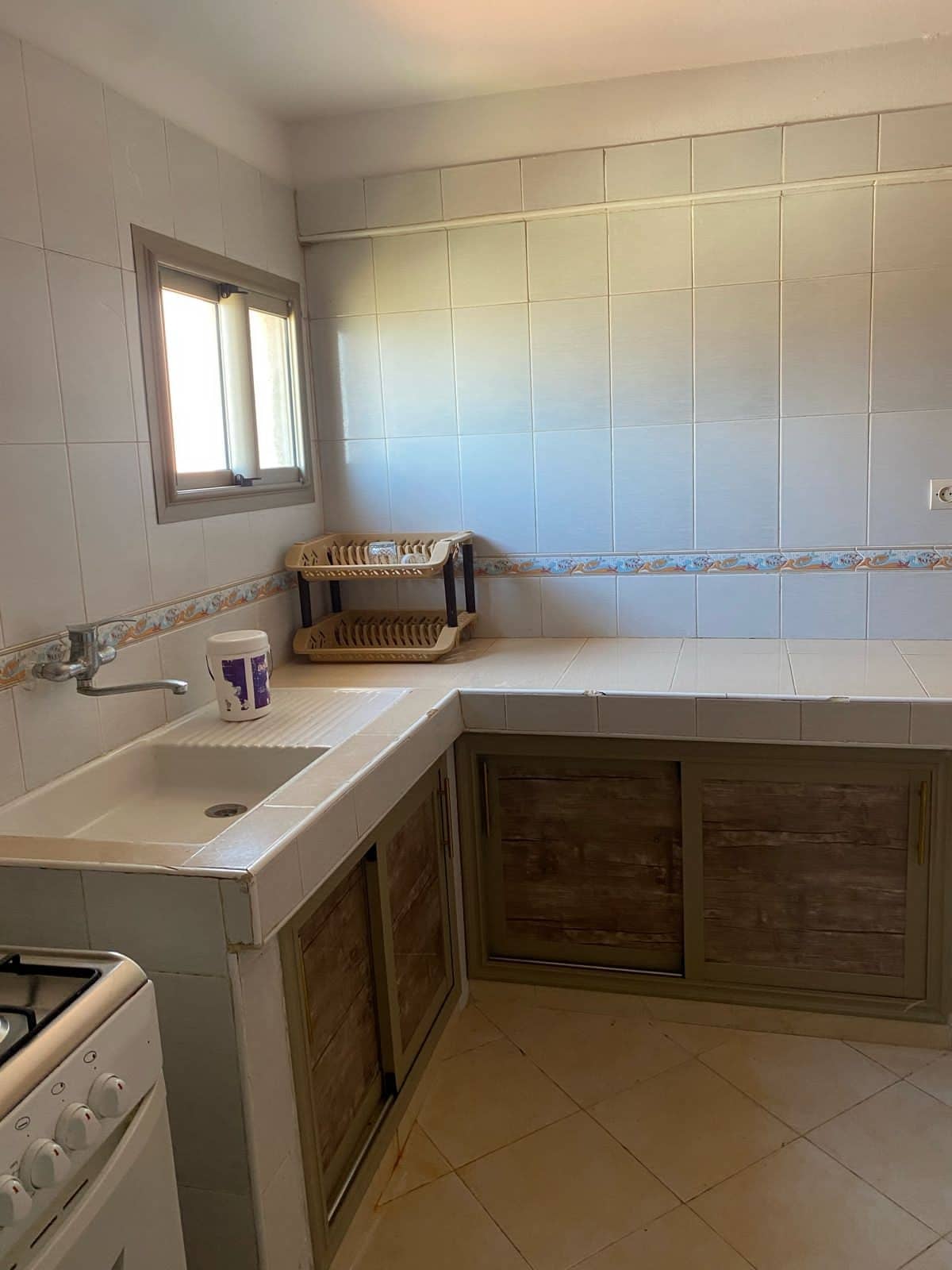 A compact kitchen features light-colored tiled walls and a window that allows natural light to enter. A sink is integrated into the countertop, accompanied by a wooden drying rack placed nearby. The space is organized with cabinetry beneath the counter.