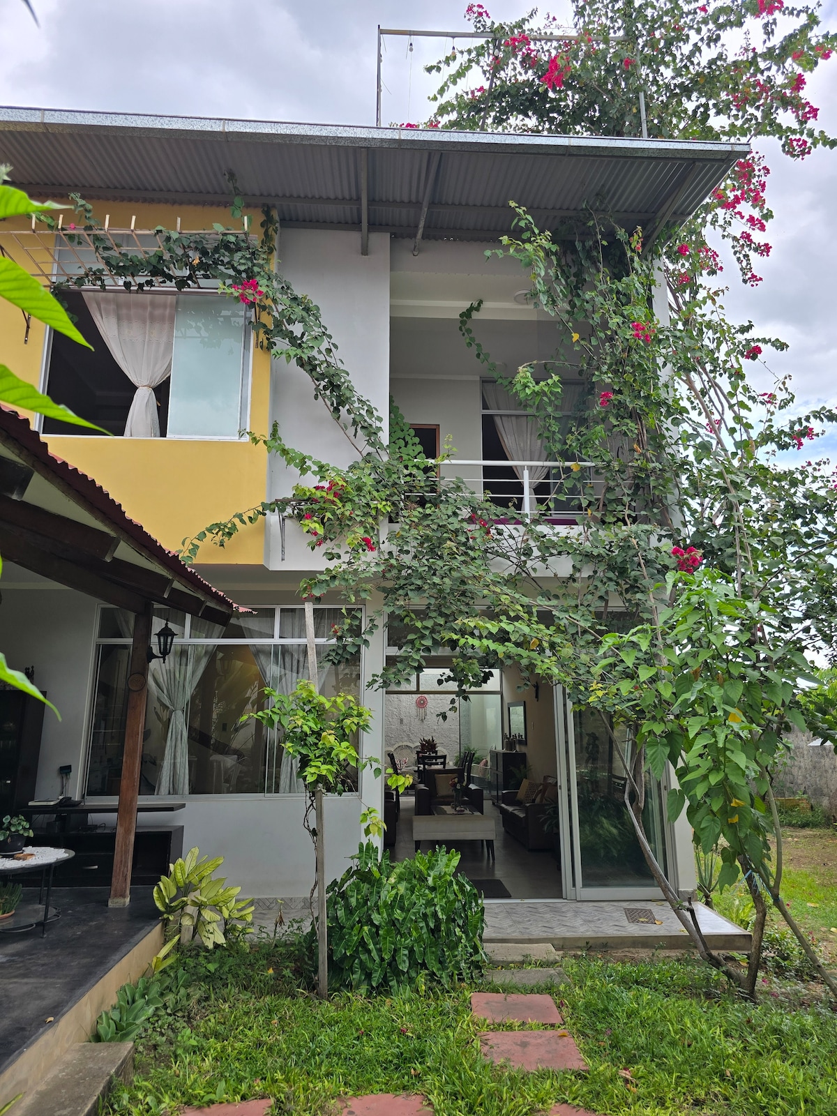 A multi-story building is surrounded by vibrant greenery, with climbing plants adorning the exterior. The facade includes large windows allowing natural light to illuminate the interiors. A staircase leads to an upper level, enhancing the vertical appeal of the structure.