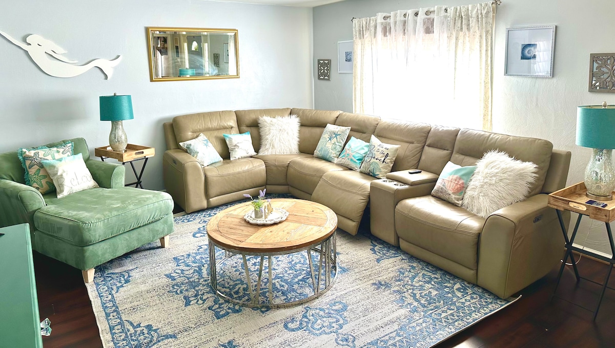 A spacious living area is furnished with a large sectional sofa, accented by decorative pillows and a cozy chair. A round wooden coffee table sits at the center, surrounded by a light blue area rug. Natural light enters through the window, framed by sheer curtains.