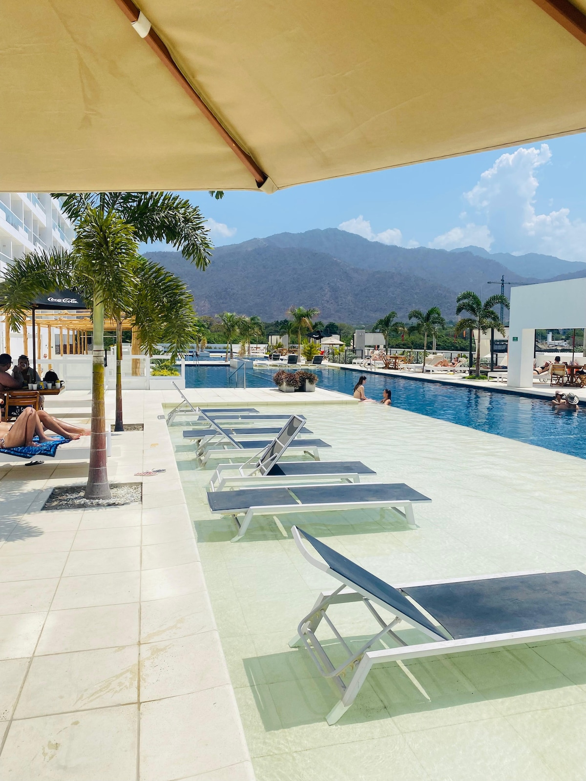 A spacious pool area is presented with several lounge chairs arranged neatly alongside the water. Lush palm trees add a touch of greenery, while distant mountains provide a scenic backdrop. The clear blue pool invites relaxation and enjoyment under the sun.