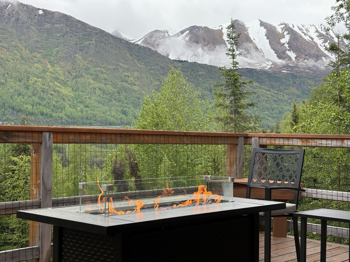 A fire pit is positioned on a wooden deck with a glass barrier, surrounded by lush greenery. Snow-capped mountains rise in the distance, under a cloudy sky, offering a serene view of the natural landscape.