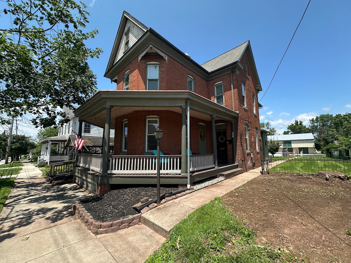 The exterior of a two-story brick home features a spacious wrap-around porch with ample seating. Lush green grass and well-maintained landscaping enhance the front yard, while a pathway leads to the entrance. The quaint neighborhood is visible in the background, accompanied by clear blue skies.