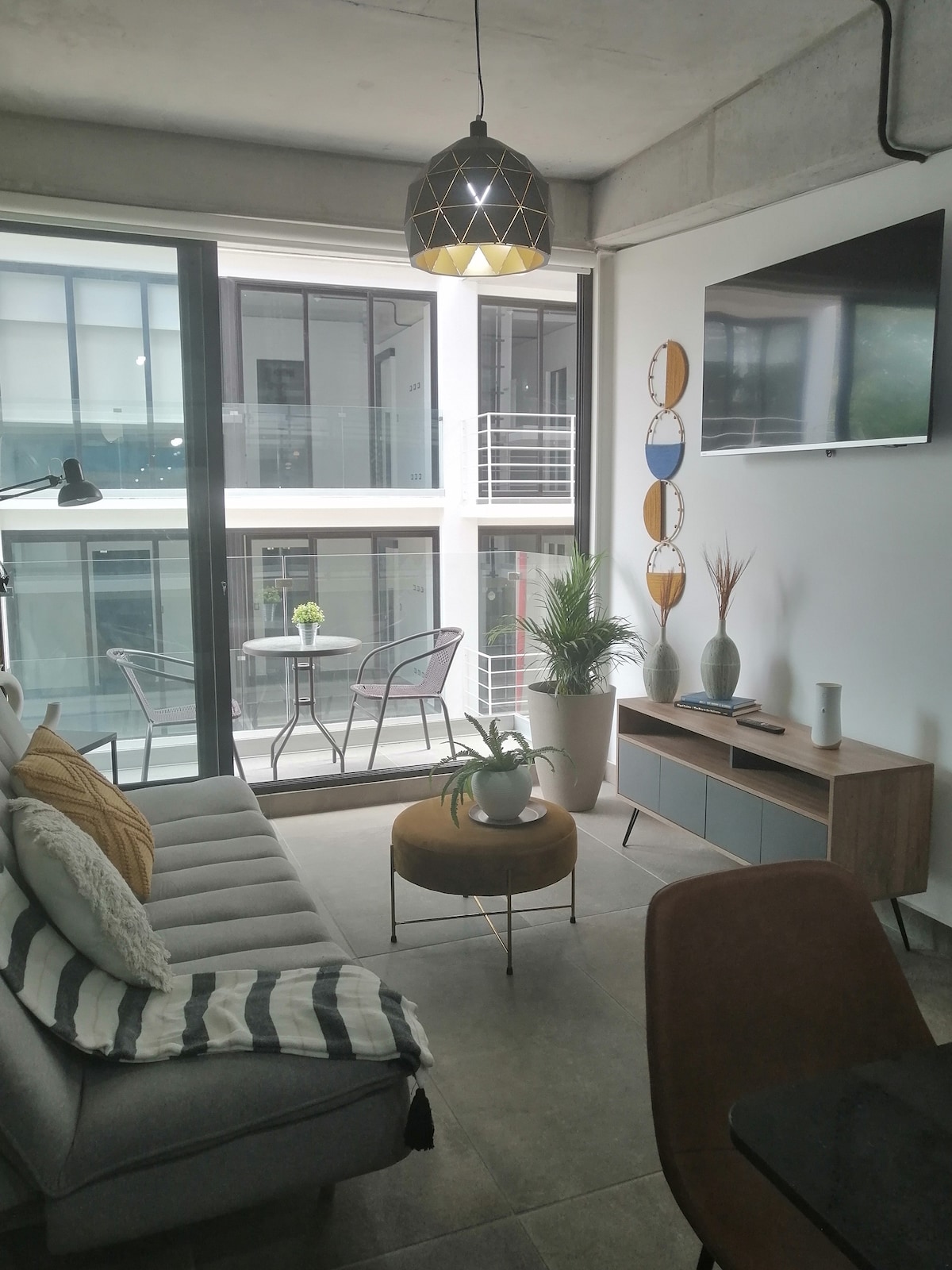 A modern living space is highlighted by large glass doors allowing natural light. A comfortable seating area features a gray sofa with decorative cushions. A television hangs on the wall above a stylish media unit. A small dining area with chairs and a table is visible by the window.