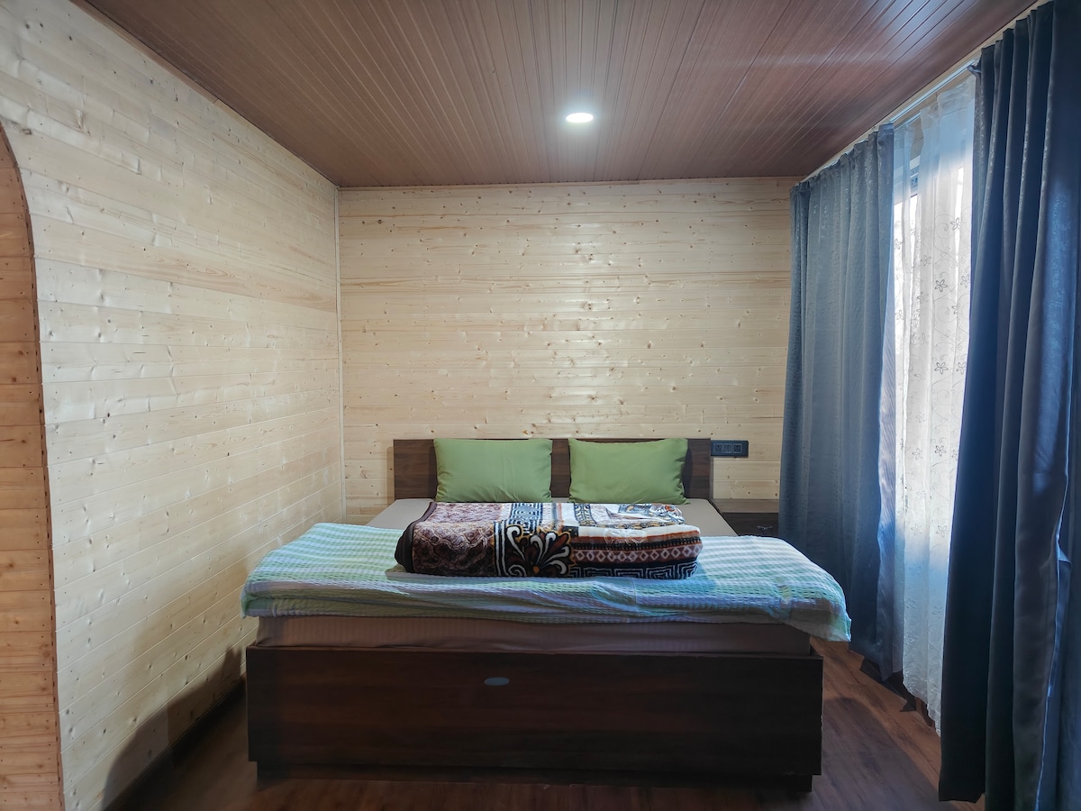 A well-lit bedroom features a spacious bed adorned with soft bedding and plush pillows. The wooden walls provide a warm backdrop, while sheer curtains allow natural light to filter through, creating a calming environment, perfect for relaxation.