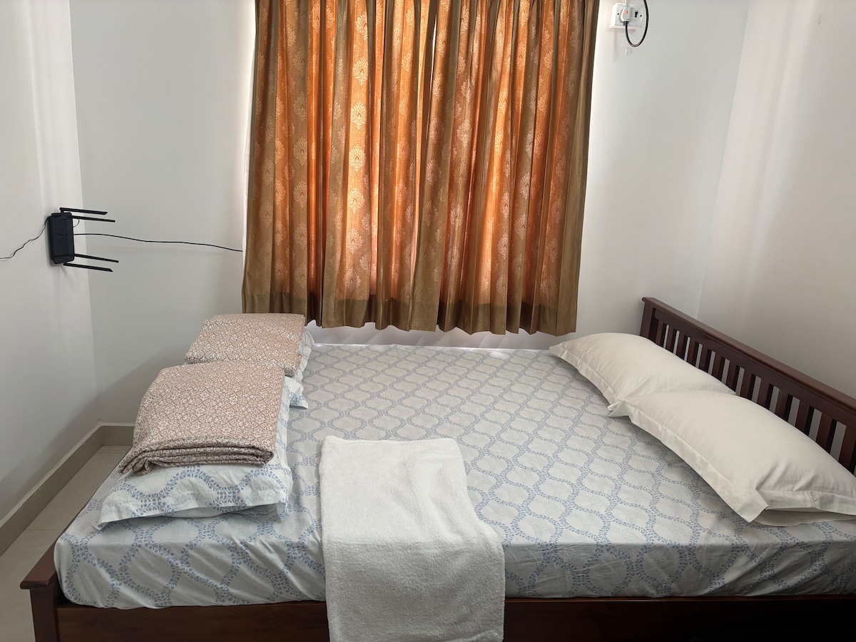 A spacious bed is layered with a patterned blanket and two white pillows. Light filters through the orange curtains, softening the room's neutral tones. A white towel is neatly placed at the foot of the bed, while a wall-mounted device is visible beside it.