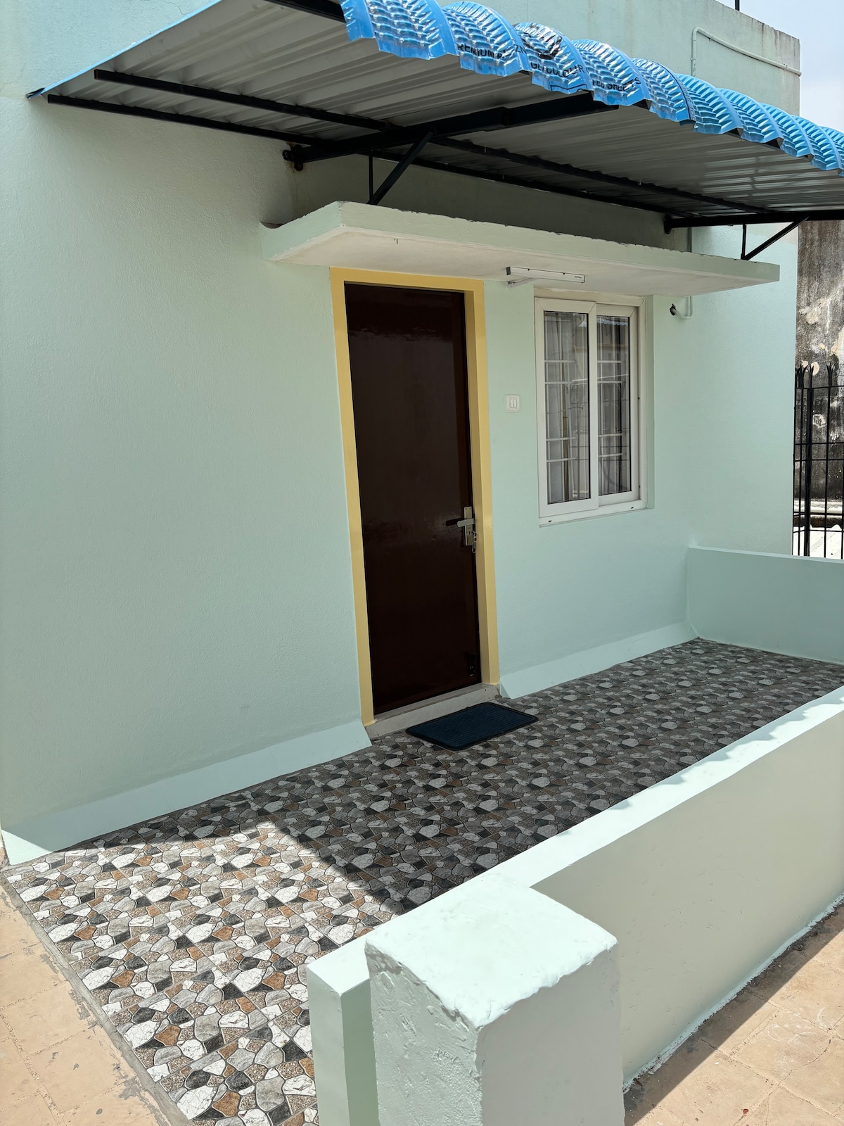 An entrance is presented, featuring a solid brown door framed by a small window. The surrounding walls are painted in light blue, and a tiled pathway leads up to the door, adding texture with a patterned design. A canopy provides shelter above the entrance.