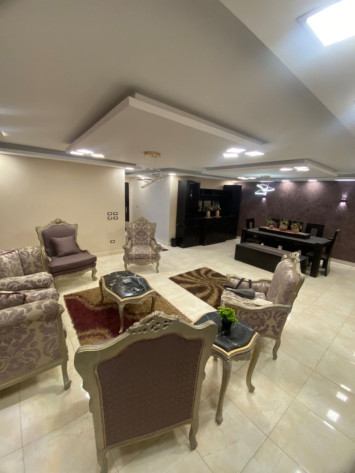 The living area is furnished with ornate chairs and a glass coffee table, complemented by a plush area rug. Soft lighting enhances the elegant design, while a dining table is visible in the background. Textured wall accents add depth to the space.