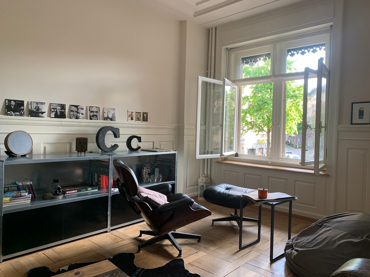 The image displays a cozy living area with a sleek black chair and a small side table. Natural light streams in through a large window featuring open panes. A glass display cabinet, adorned with books and decorative items, sits against the wall. Greenery is visible outside.