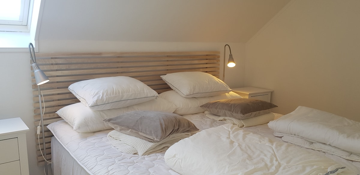 A spacious sleeping area is presented with a wooden headboard featuring multiple fluffy pillows layered atop a large bed. Soft lighting is provided by two bedside lamps, while a nightstand is positioned beside the bed. The room is well-lit with natural light streaming through a nearby skylight.