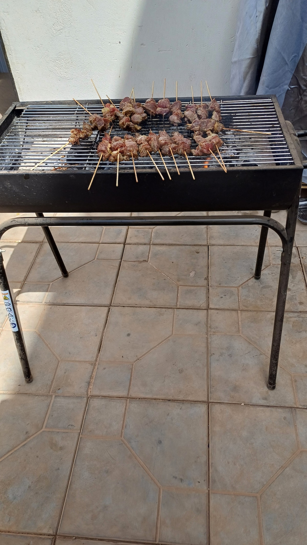 A barbecue grill sits on a sturdy metal frame, filled with skewers of marinated meat. The grill is positioned on a tiled surface, with a clean and organized appearance, ensuring a functional outdoor cooking space.