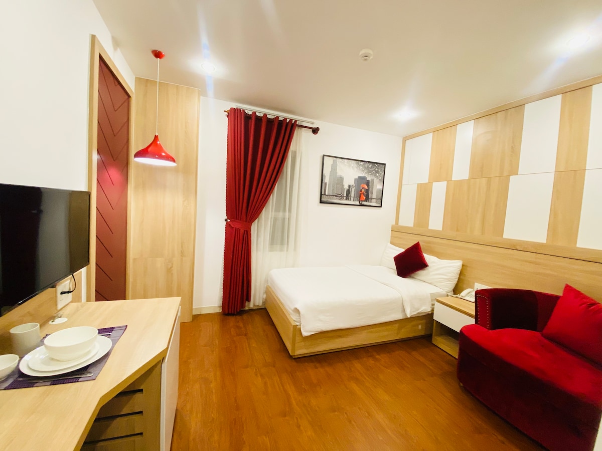 A serene guest room features a single bed dressed in crisp linens, complemented by a plush armchair and a wooden bedside table. Natural light enters through the window, while rich red curtains add a pop of color. A modern television is mounted on the wall.