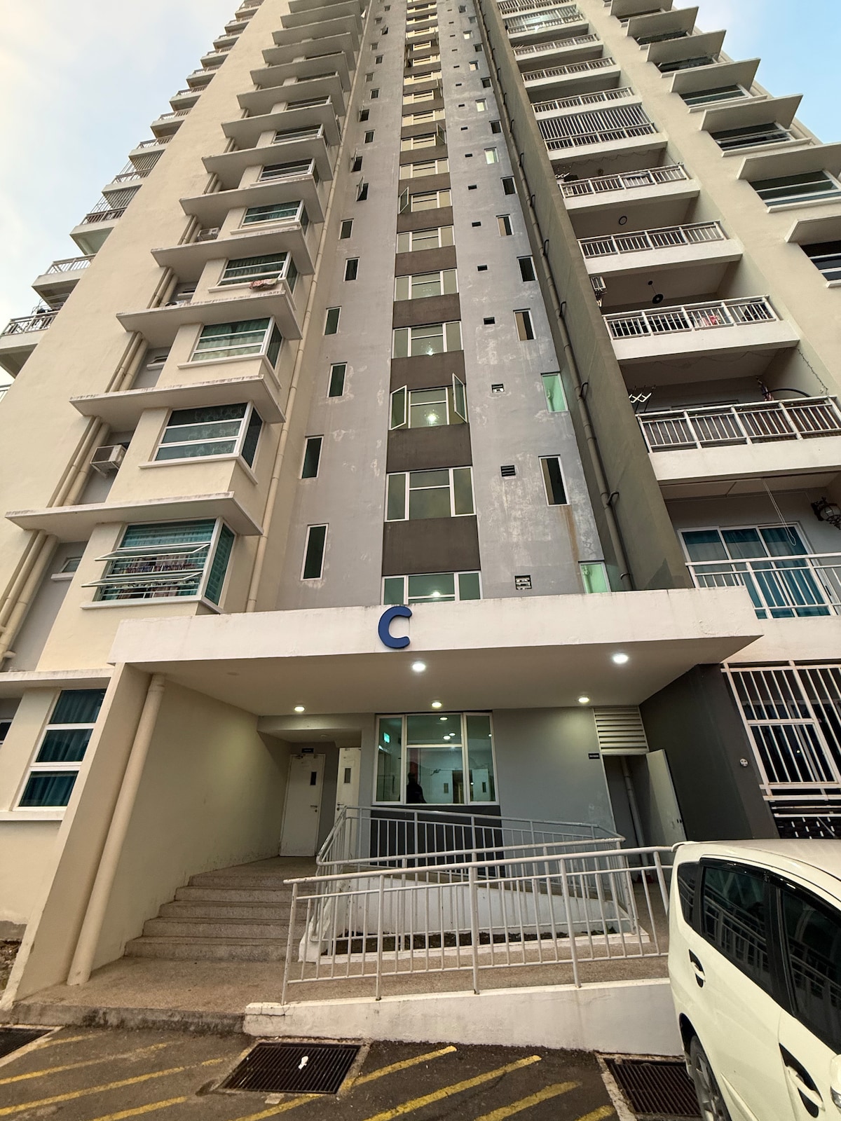 The exterior of a tall building is featured, showcasing a modern design with multiple floors and large windows. An entrance with steps and a ramp is visible, promoting accessibility. The surrounding area is organized with parking spaces for vehicles.