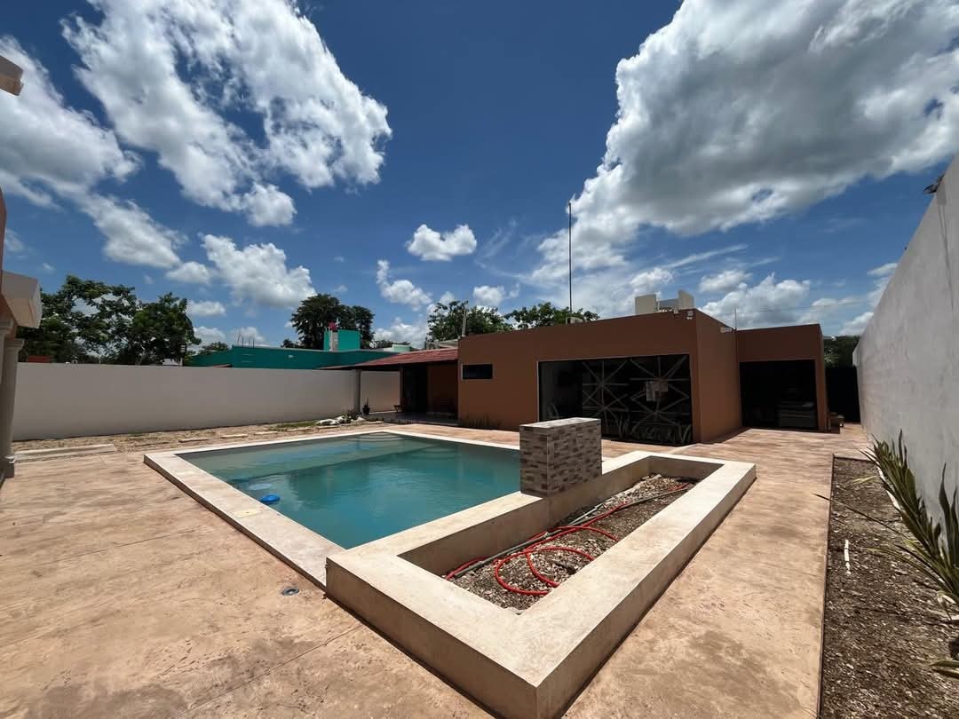 A spacious outdoor area features a rectangular swimming pool, surrounded by a clean, concrete deck. A single structure is visible, showcasing large glass doors and a modern aesthetic. Lush greenery is present in the background, under a partly cloudy sky.