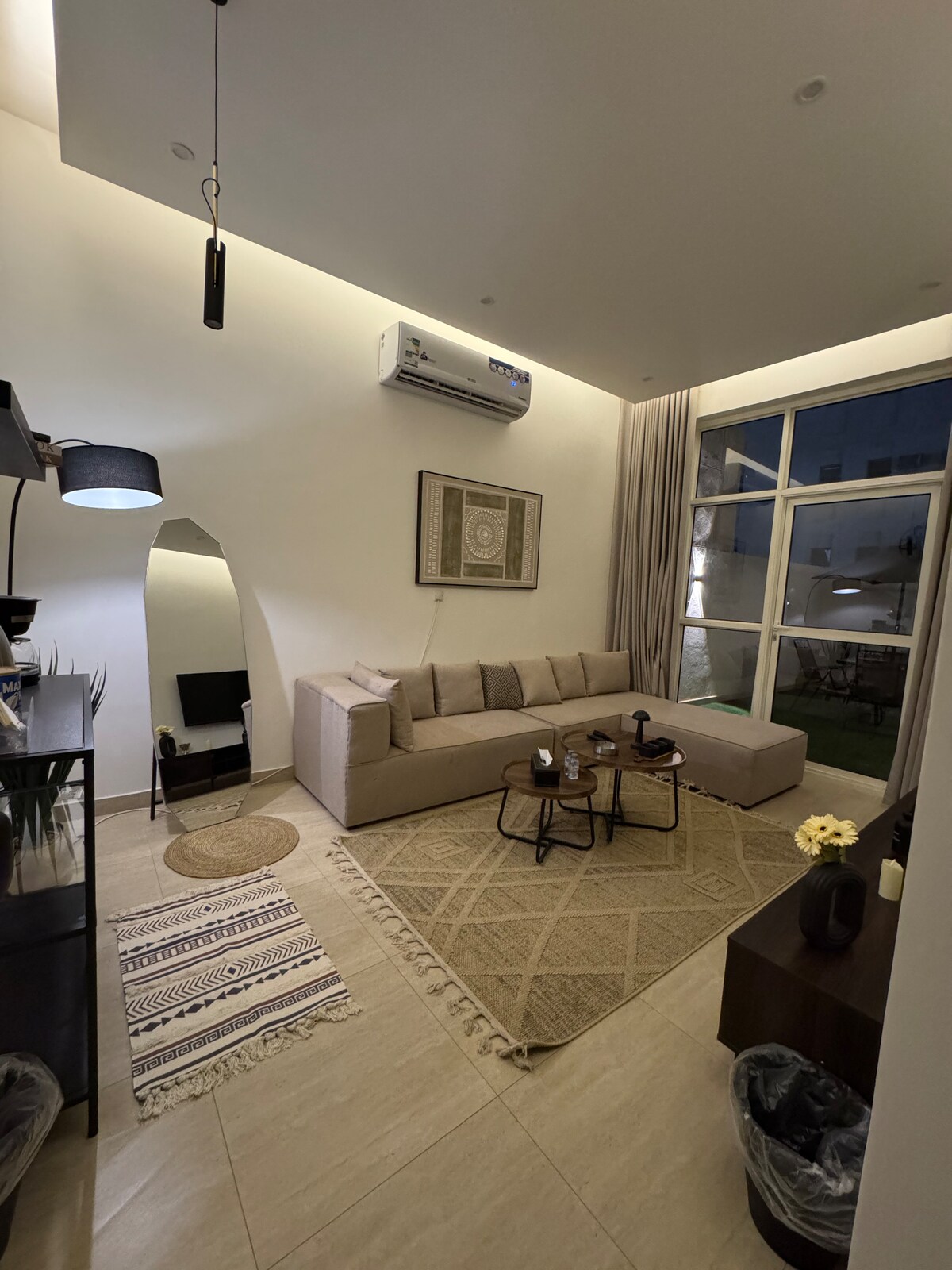 A spacious living area is showcased with a comfortable sectional sofa arranged around a pair of coffee tables. Natural light flows through large windows, illuminating the room. A decorative rug lays on the floor, and a small TV is positioned on a nearby cabinet.