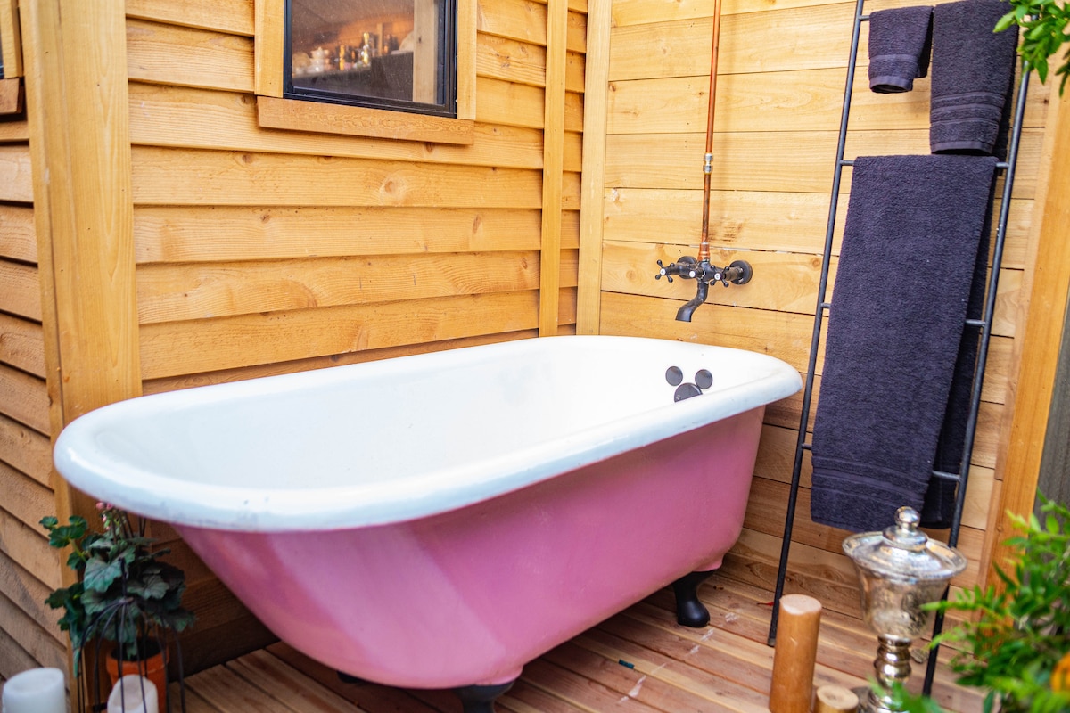 An antique clawfoot tub with a pink exterior is positioned between wooden walls, evoking a sense of outdoor relaxation. A showerhead is mounted above, and two dark towels hang on a rack nearby. Potted plants and decorative items add subtle charm to the rustic ambiance.