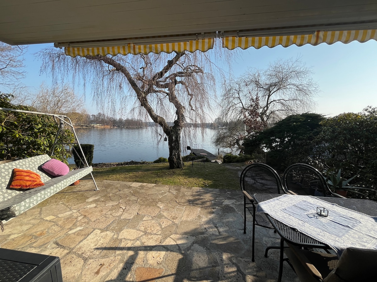 A stone patio offers a serene view of the lake, framed by leafy trees. A swing chair is positioned nearby, adorned with colorful cushions. A table with chairs is set for outdoor dining, inviting relaxation and enjoyment of the natural surroundings.