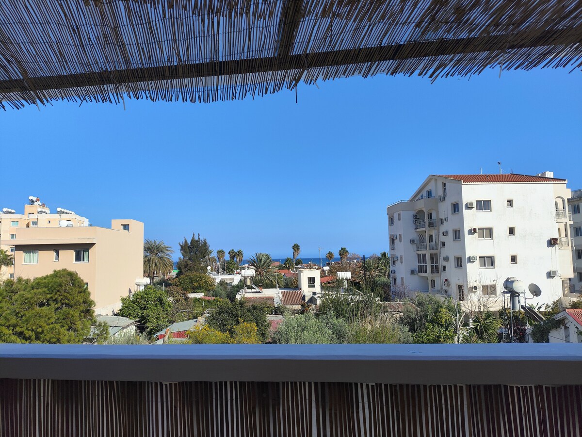 An expansive view reveals a clear blue sky overlooking a landscape of palm trees and buildings. The image captures the sea in the distance, framed by the natural texture of a bamboo covering, providing a serene backdrop.