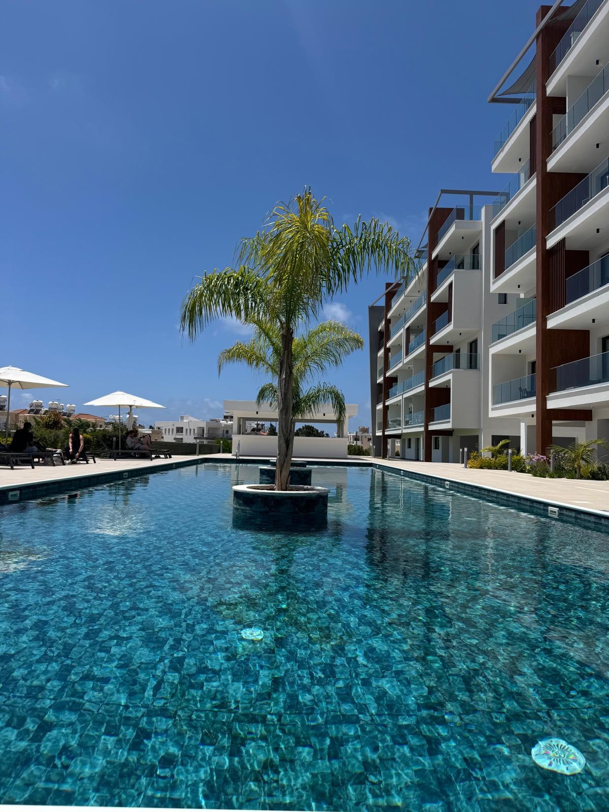 A serene pool area features a central palm tree surrounded by clear, sparkling water. White umbrellas are positioned around the poolside, and modern residential buildings are visible in the background, reflecting a tranquil atmosphere suitable for relaxation.