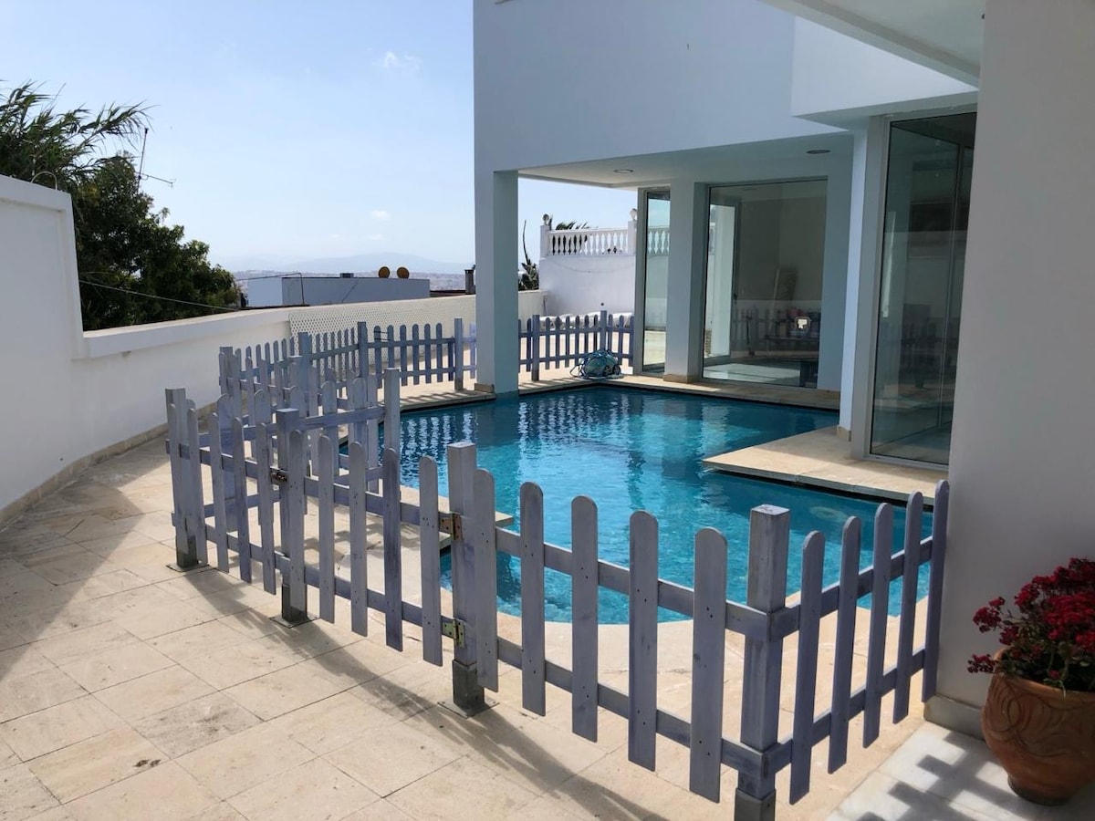 A private pool is framed by a light blue wooden fence, providing a secure environment. The pool, reflecting clear water, is surrounded by a spacious stone patio, with a few potted plants visible. The backdrop features a partially open area leading to the villa's interior.