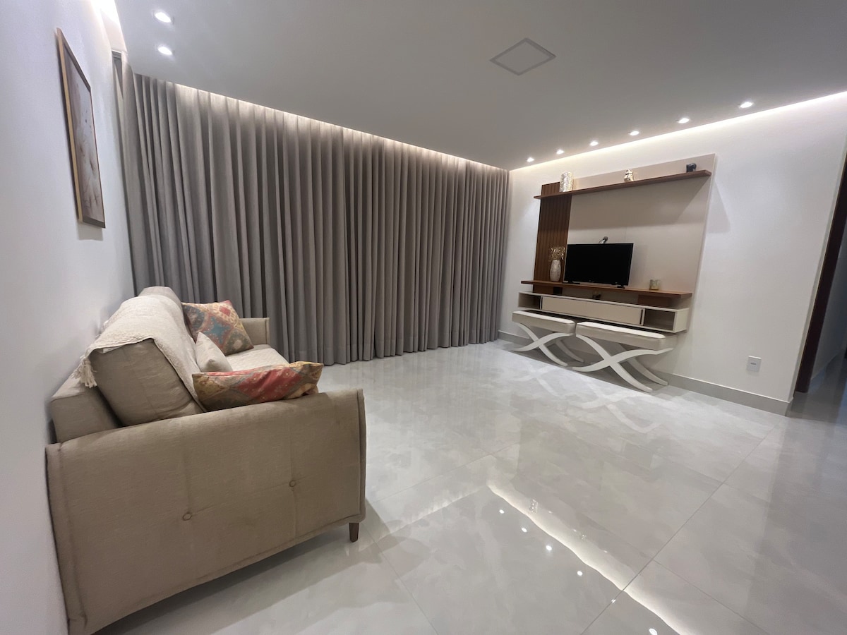 A spacious living area features a light-colored sofa adorned with decorative cushions. Soft, sheer curtains frame the large window, allowing natural light to enhance the glossy tile floor. A modern entertainment unit with a TV occupies one wall, complemented by subtle ambient lighting.