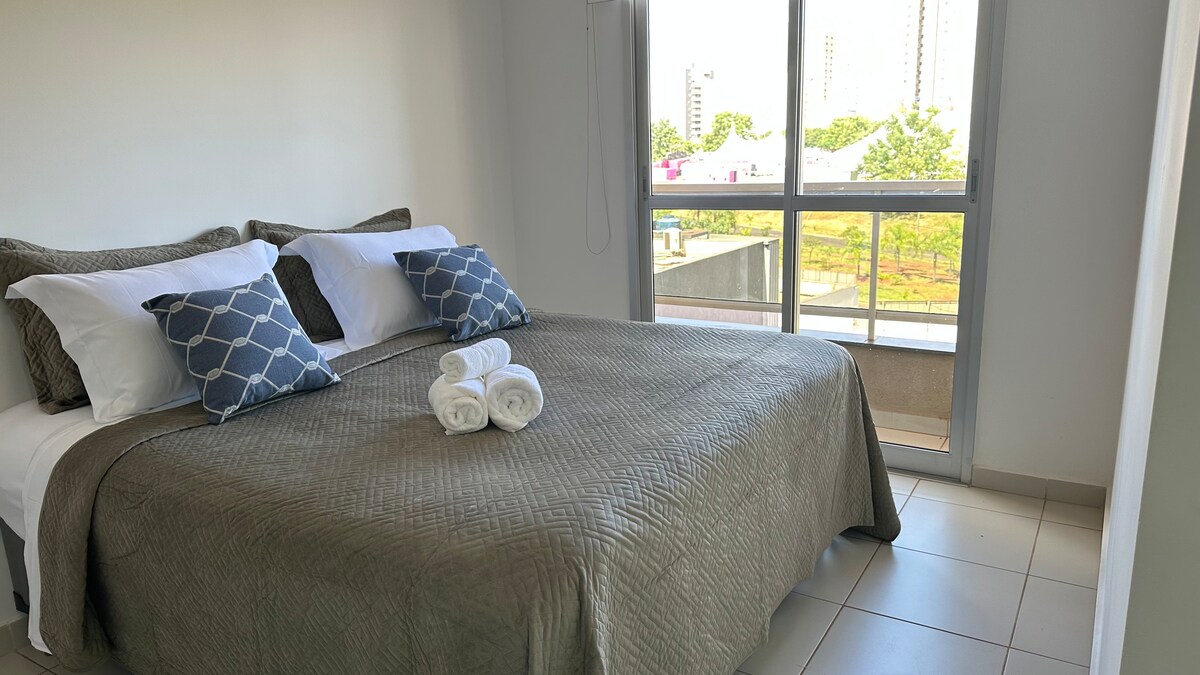 A spacious sleeping area features a soft, neutral-toned bedspread covering a large bed adorned with two patterned cushions. Fresh white towels are neatly arranged at the foot of the bed. Natural light enters through a nearby window, overlooking greenery.