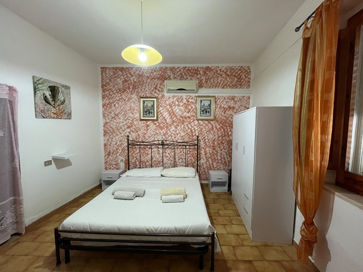 A bedroom featuring a metal bed frame with an unmade mattress, accompanied by two neatly folded towels. The room is adorned with a patterned accent wall and two framed artworks. A white wardrobe stands against the wall, with soft lighting provided by a ceiling fixture.