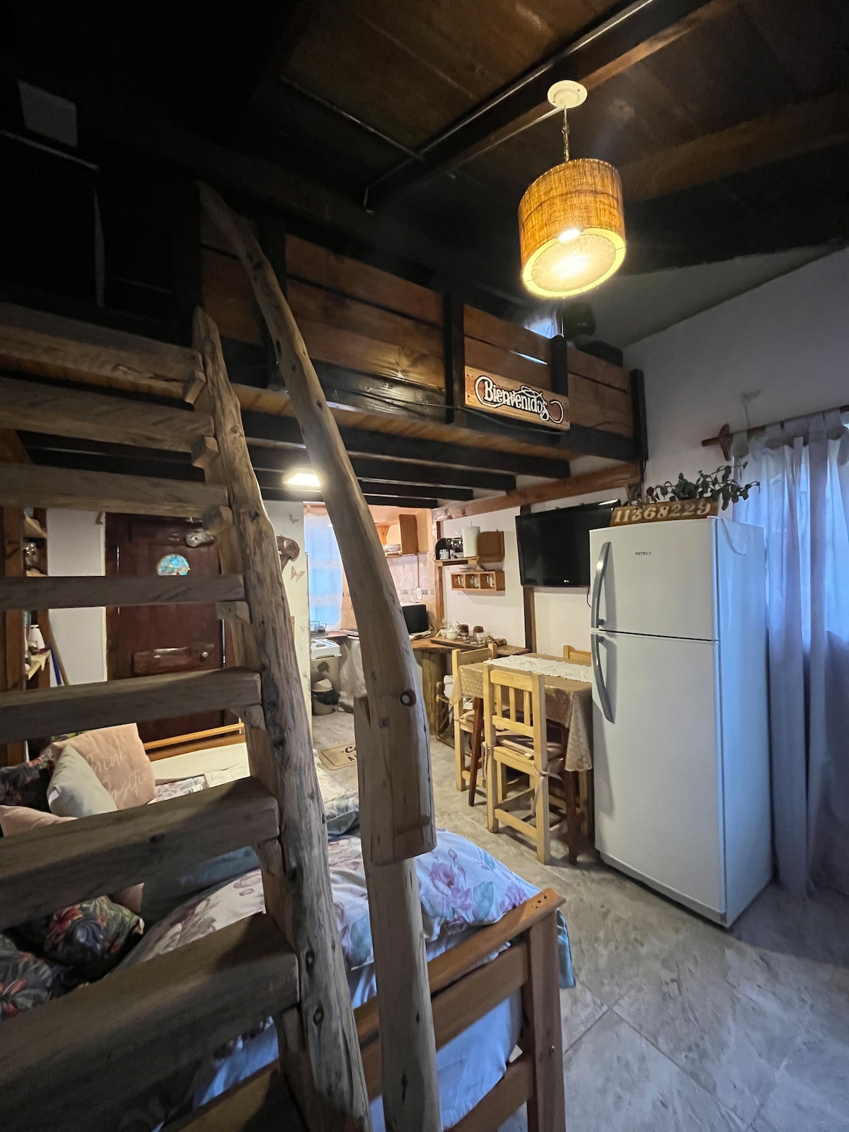 A wooden staircase leads to a lofted area in a cozy living space. The room features an open kitchen with a white refrigerator and a small dining table with stools. Natural light enters through a window, illuminating floral-patterned furnishings.