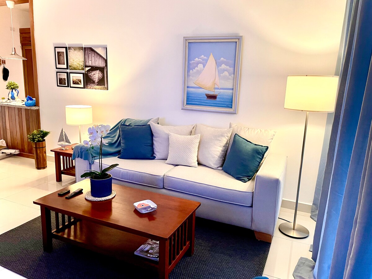 A bright living room is featured, showcasing a white sofa adorned with blue and white cushions. A wooden coffee table rests in front, accompanied by a potted plant. A simple painting of a sailboat is displayed on the wall, and two floor lamps provide gentle illumination.
