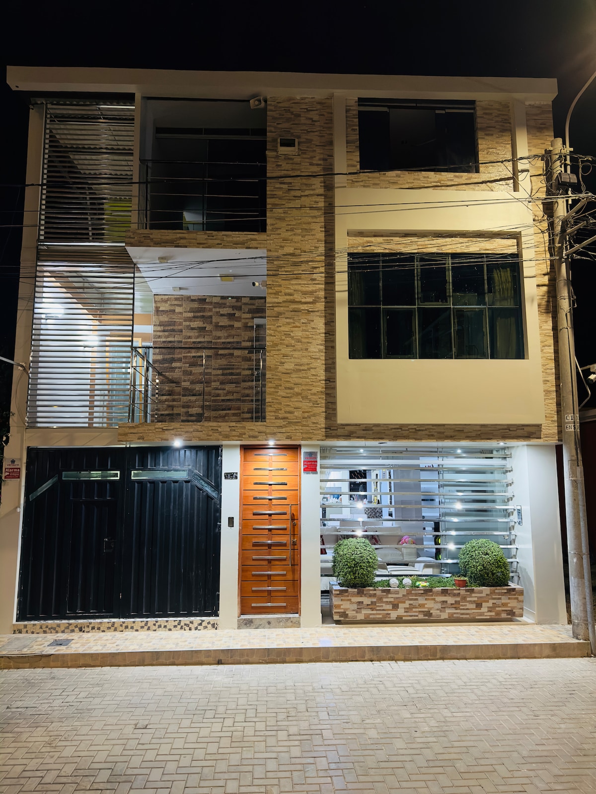 The exterior facade of a three-story building is showcased at night, illuminated by soft lighting. The entrance features a modern wooden door, complemented by large glass windows. Decorative plants are positioned symmetrically on either side of the entrance, enhancing the building's sleek design.