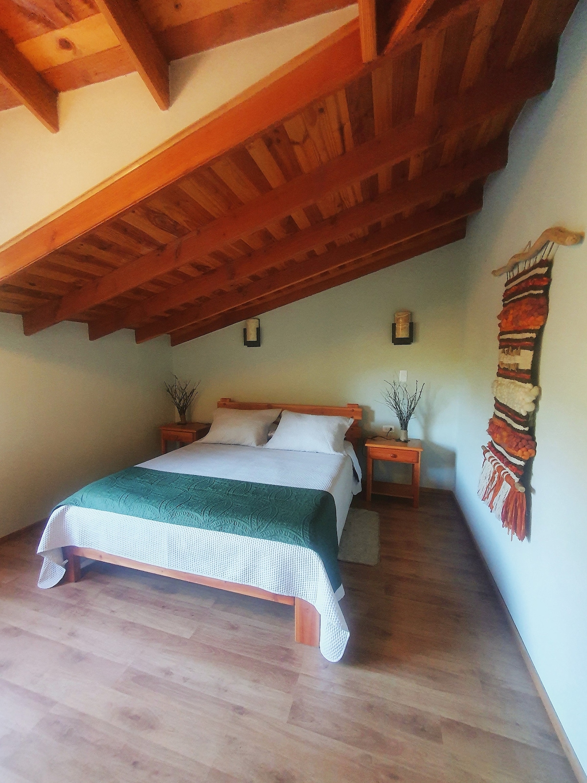 A bedroom features a spacious bed covered with a green blanket, complemented by light-colored sheets. Two wooden nightstands are positioned on either side of the bed. The ceiling showcases exposed wooden beams, and a decorative wall hanging adds a touch of warmth to the room.