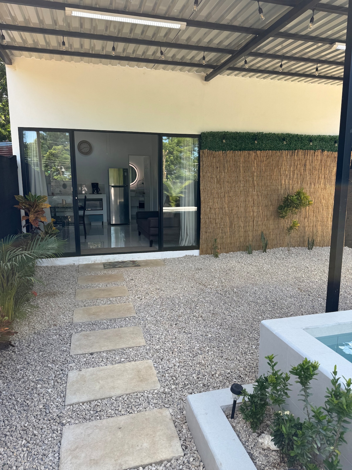 A modern exterior view showcases a minimalist villa with large glass sliding doors opening to a tranquil interior. Stone pathways wind through a gravelled yard adorned with greenery, leading towards the entrance. Natural elements are incorporated into the landscape, creating a serene outdoor atmosphere.