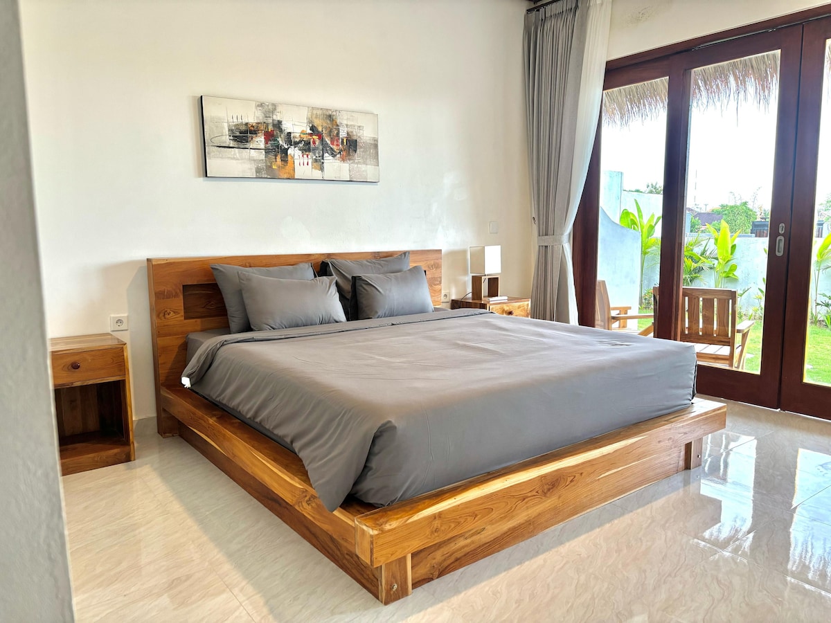 A spacious bed with neutral grey bedding is set against a wooden frame. Two bedside tables flank the bed, each with a lamp. Large glass doors open to a green patio area, allowing natural light to brighten the room.