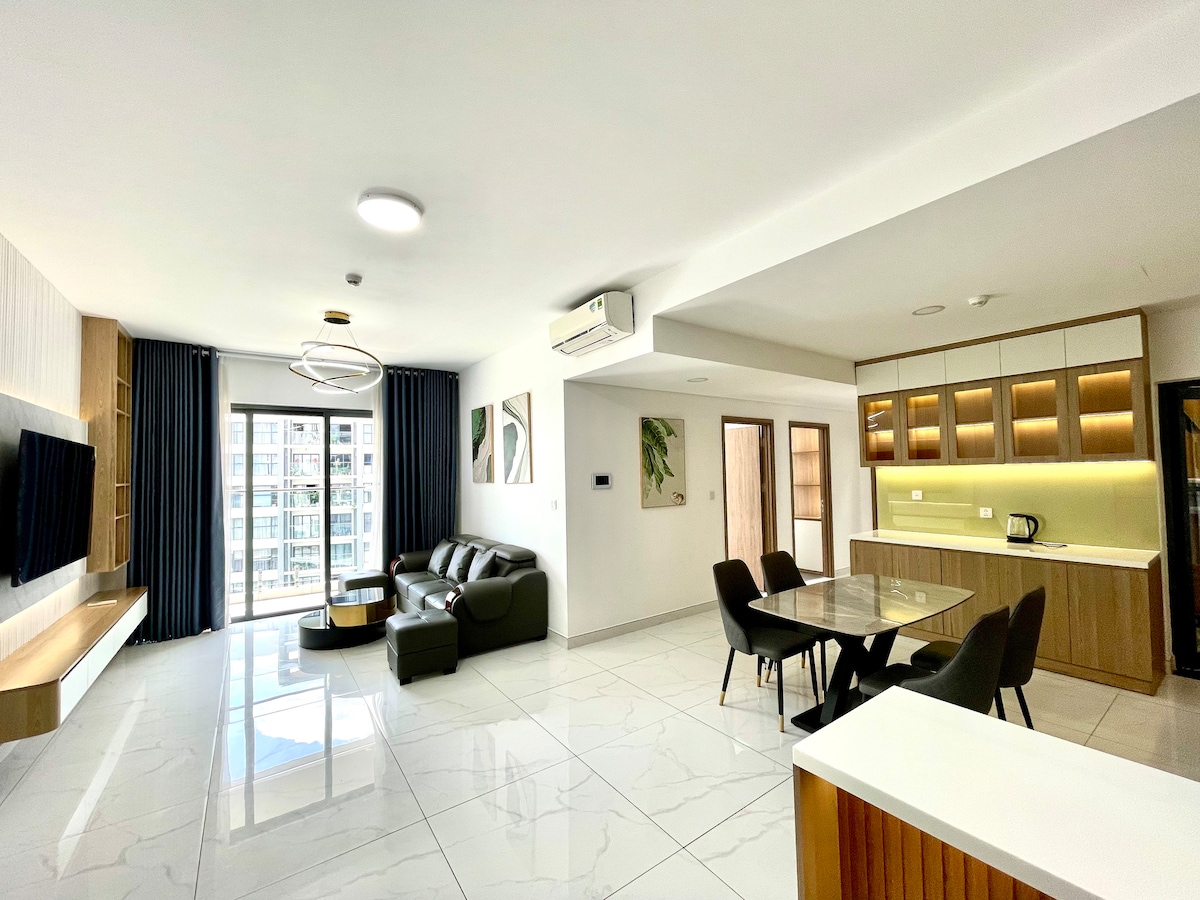 A spacious living area features a dark leather sofa arranged around a glass coffee table. A dining area with a round table is visible, accompanied by a sleek kitchen in the background. Large windows allow ample natural light to fill the space.