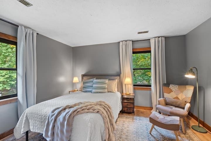 Chumba cha kulala cha kifalme chenye mwanga na utulivu chenye rangi laini za asili, matandiko ya kifahari na kiti cha starehe kinachofaa kwa kusoma au kupumzika.