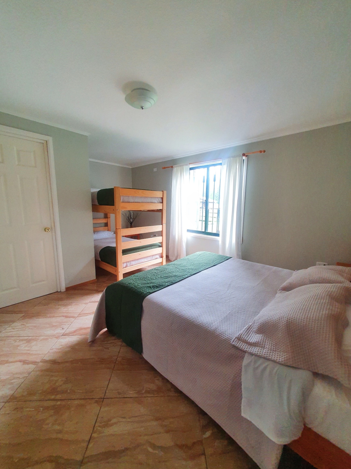 A bright sleeping area is designed with light-colored walls and tiled floors. A spacious bed is accompanied by a set of bunk beds nearby. Natural light enters through a window, enhancing the cozy environment, while soft linens in neutral tones are arranged on the beds.