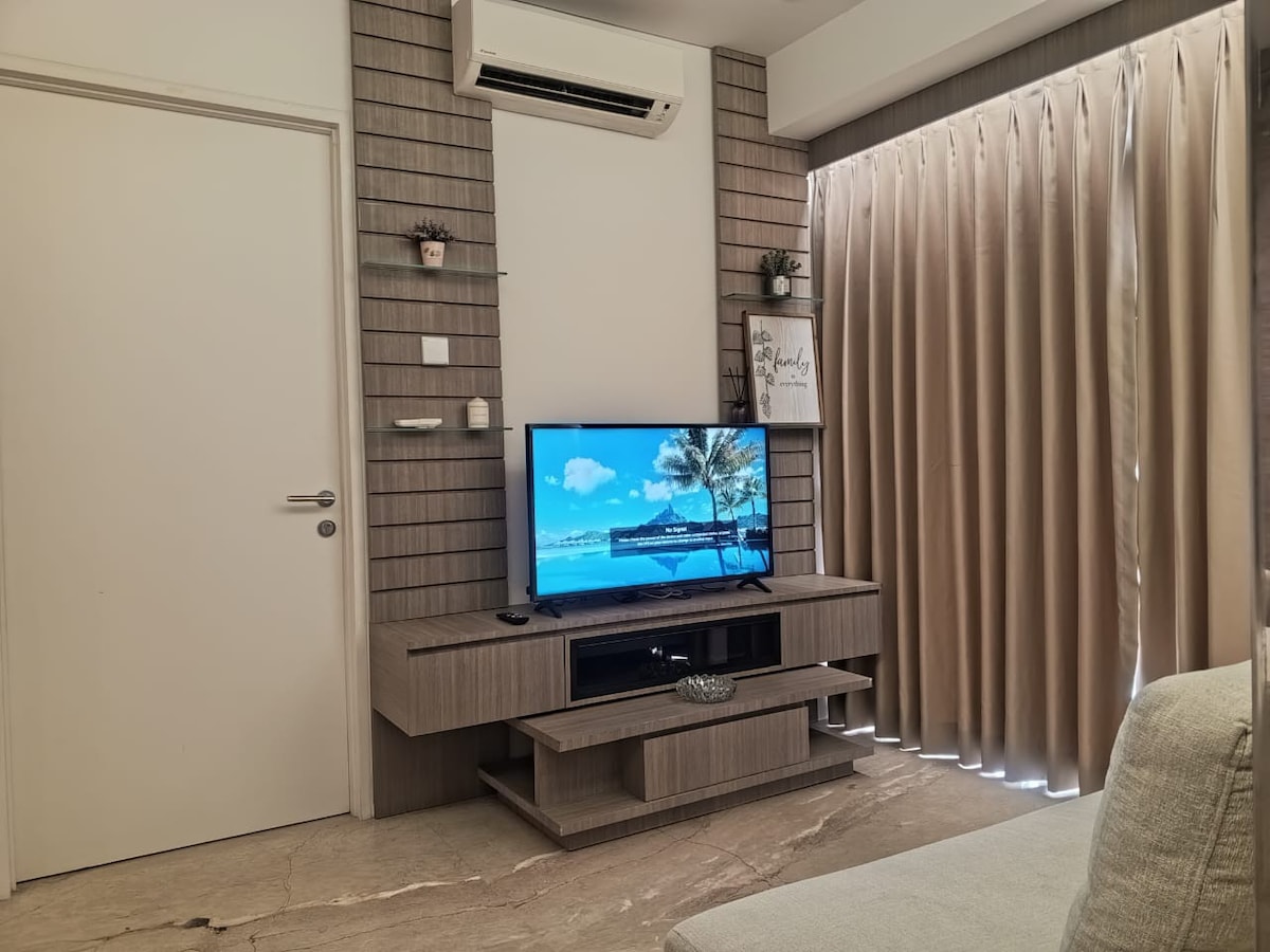 A modern living area features a wall-mounted television above a sleek entertainment unit. Neutral-colored curtains partially cover the window, allowing soft light to fill the room. An air conditioning unit is positioned above the door, providing comfort during warm days.