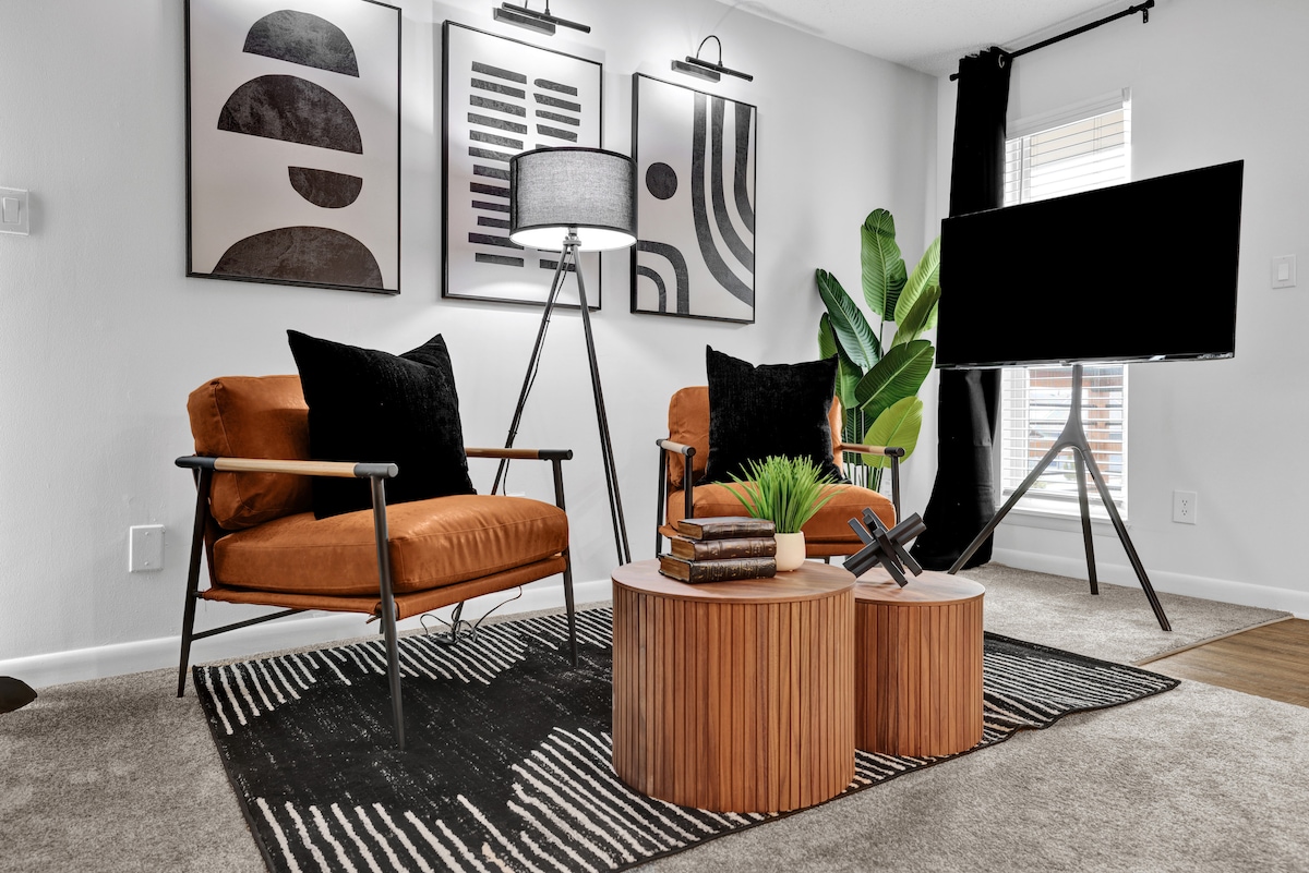 A cozy seating area features two orange chairs with black cushions, positioned beside a wood coffee table. Decorative plant foliage is present, and modern artwork adorns the walls. A standing lamp and a black television on a tripod stand complete the stylish layout.