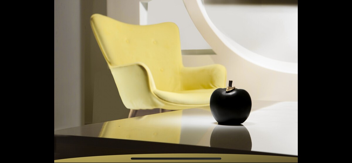 A soft yellow chair is positioned nearby a sleek surface, displaying a black decorative apple with a subtle shine. The light reflects gently off the smooth table, creating a modern and stylish ambiance.