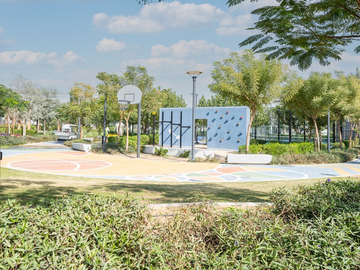 An outdoor recreational area is displayed, featuring a basketball hoop alongside a vibrant painted surface. Lush greenery surrounds the area, with shrubs and trees providing shade. The scene is complemented by a bright sky and scattered clouds, enhancing the inviting atmosphere for play.