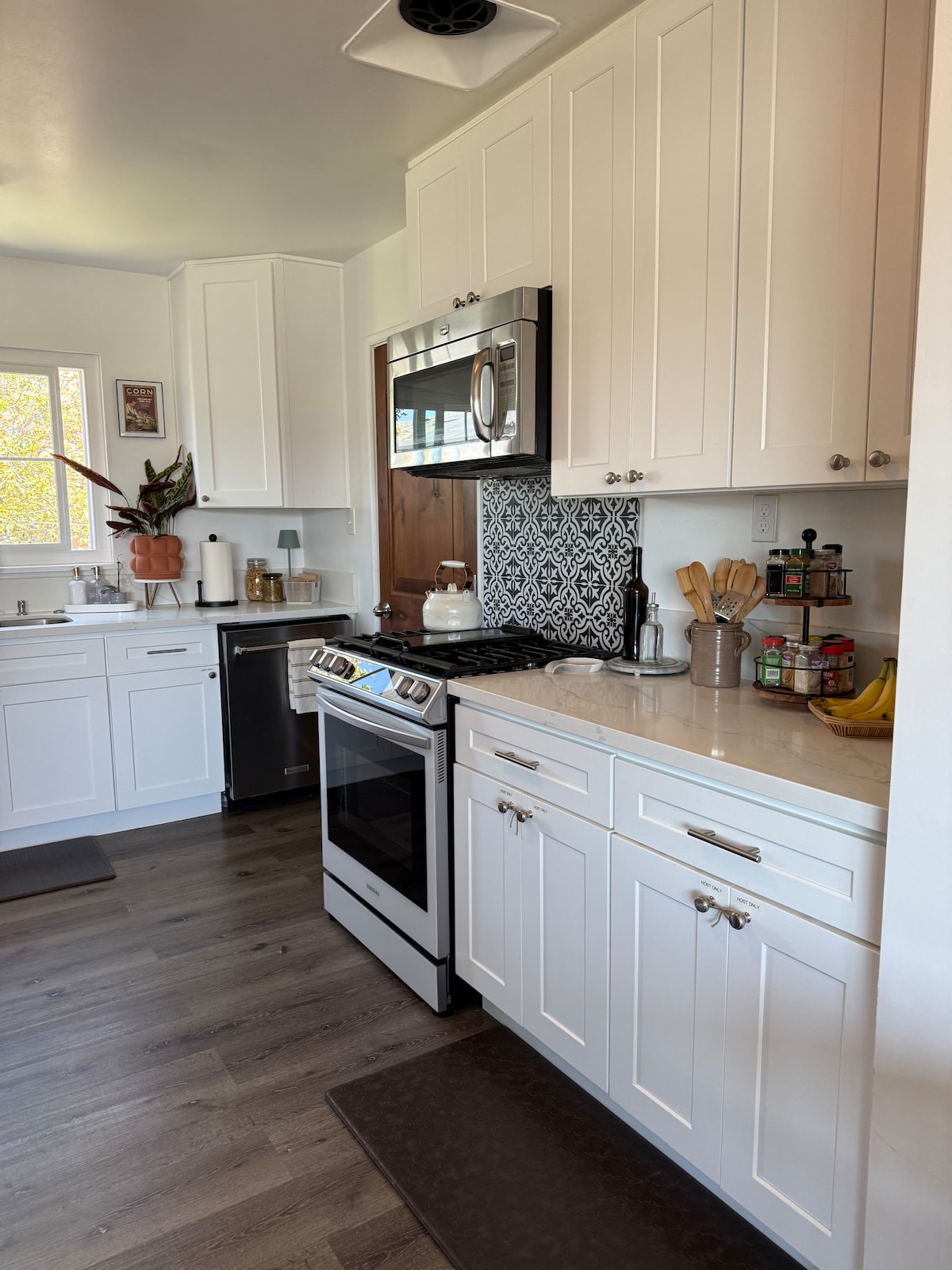 The kitchen features modern white cabinetry and a sleek stainless steel oven. A decorative backsplash adds a touch of style, while various kitchen tools are neatly arranged on the counter. Natural light fills the space through a window, creating an inviting cooking area.