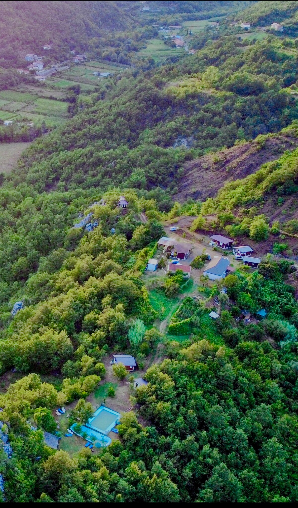 An aerial view captures a serene countryside landscape, showcasing a cluster of accommodations nestled among lush green trees. A swimming pool is visible, surrounded by open space, while the mountains frame the background, enhancing the tranquil setting of this rural retreat.