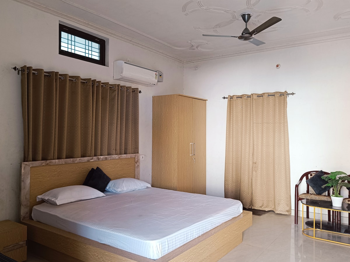 The spacious bedroom features a large bed with a neutral-colored headboard. Light-colored curtains frame a window, and a ceiling fan is mounted above. A wooden wardrobe stands against the wall, complemented by an accent chair adorned with greenery.