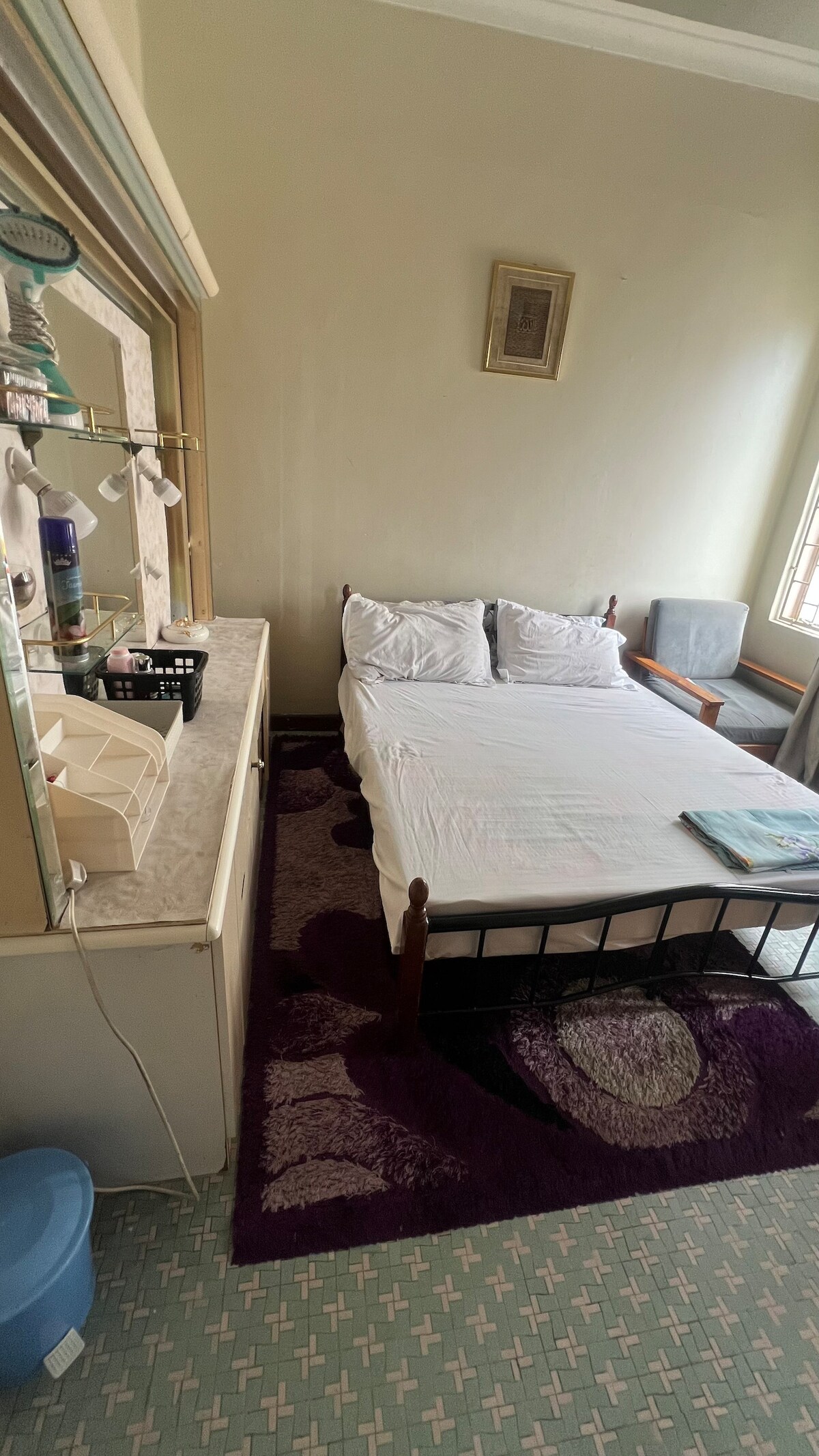 A small room is shown, featuring a single bed with white linens and pillows. A wooden dresser is positioned against one wall, while a gray armchair adds seating. A colorful rug covers the floor, and natural light enters through a nearby window.