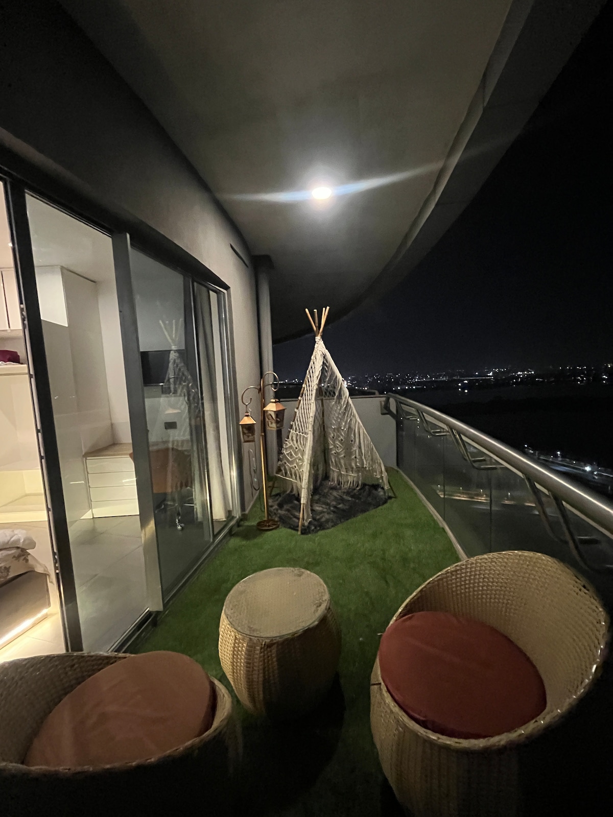 A private balcony is illuminated softly by a ceiling light. A cozy tent-like structure is positioned beside the glass railing. Two round outdoor chairs with cushions are available, enhancing the space while the city skyline is visible in the distance at night.