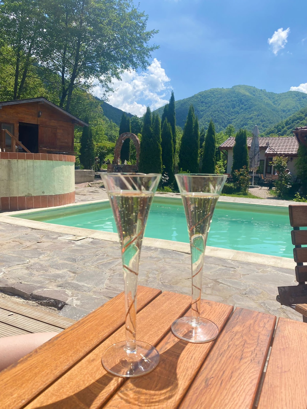 Two elegant glasses are placed on a wooden table, positioned near a clear swimming pool surrounded by green trees. In the background, mountains create a serene backdrop, while a charming wooden structure is partially visible.