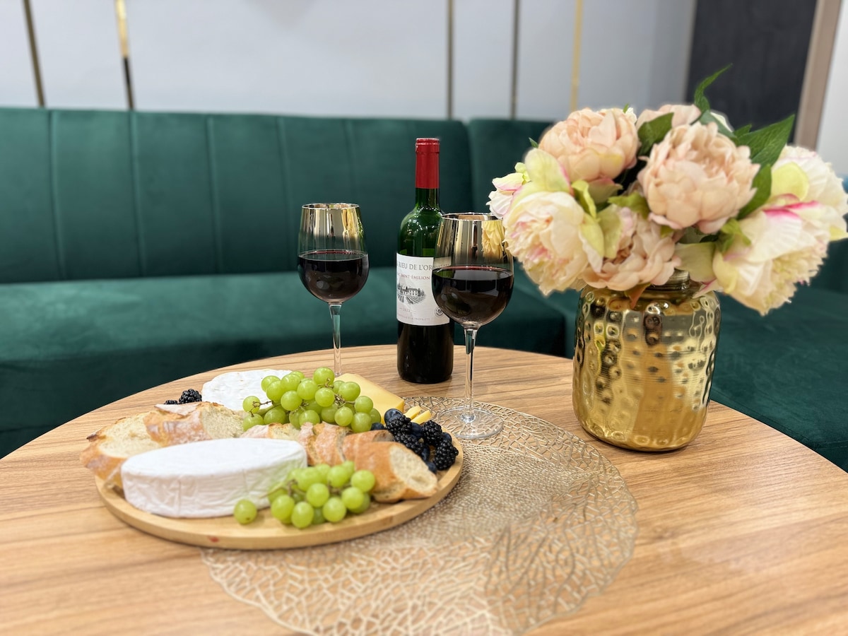 An elegant coffee table adorned with a wooden platter featuring a selection of cheeses, grapes, and croissants. Two glasses filled with red wine sit adjacent to a decorative vase of soft-colored flowers, adding a touch of sophistication to the setting.